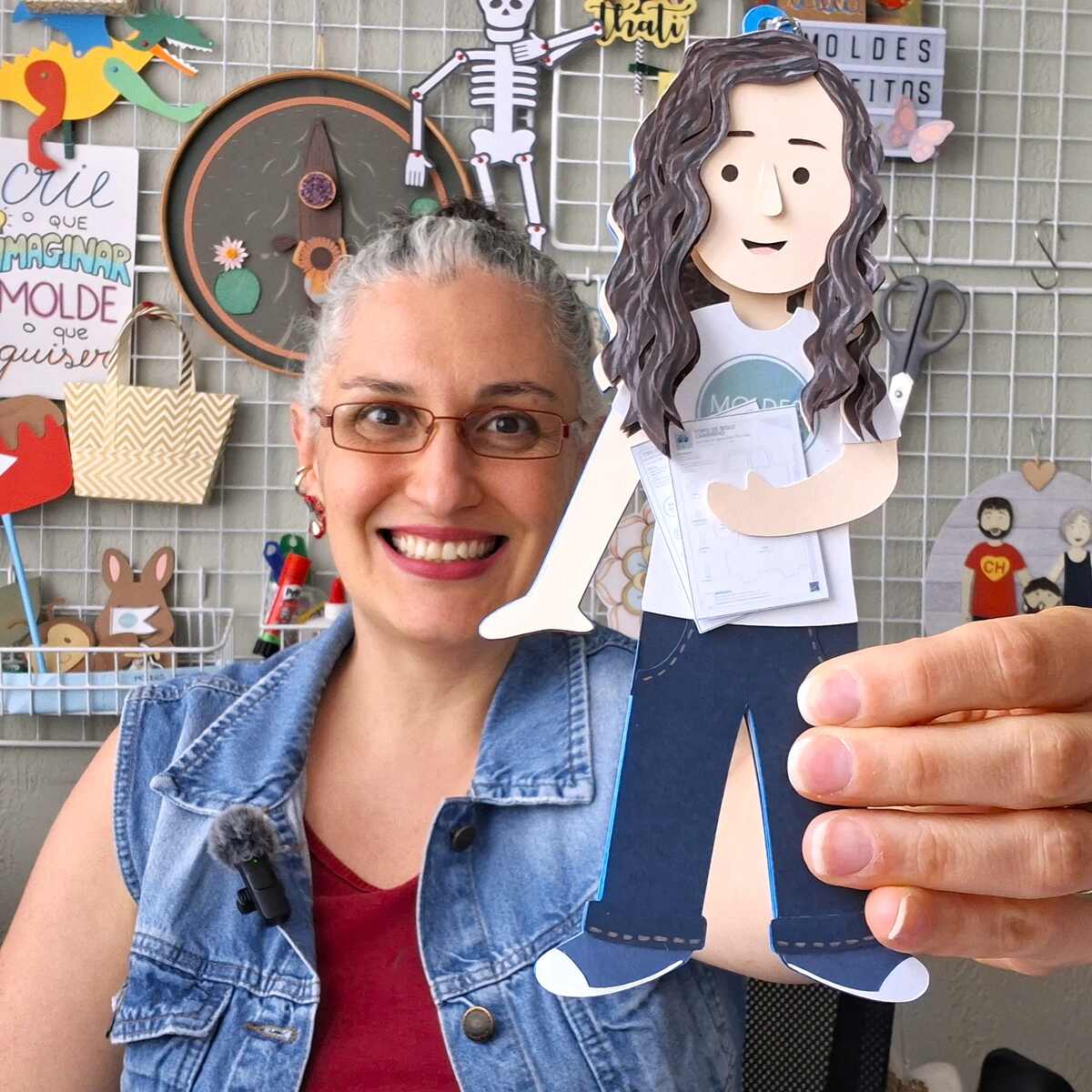 Thati Dias em seu ateliê, sorrindo e segurando uma boneca de papel artesanal
